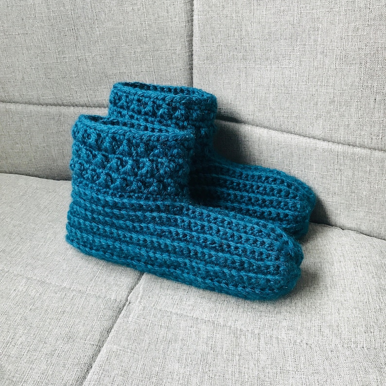 K&ouml;nnte beinhalten: Ein Paar tealblaue geh&auml;kelte Hausschuhe. Die Hausschuhe sind mit einem strukturierten Stich gearbeitet und haben ein weiches, gem&uuml;tliches Aussehen.