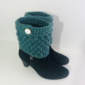 May include: A pair of black suede boots with a green crocheted cuff and a white button. The boots have a block heel and a zipper on the side.