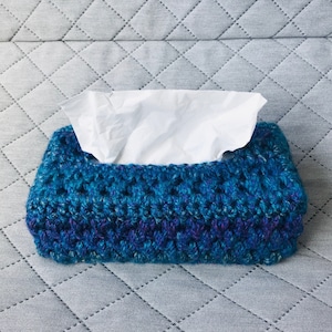 May include: A crocheted tissue box cover in shades of blue and purple. The cover is made with a textured stitch and has a rectangular shape.
