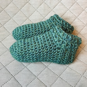 Può includere: Un paio di pantofole verdi all'uncinetto. Le pantofole sono realizzate con un punto testurizzato e hanno un aspetto morbido e accogliente.