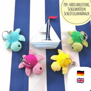 Peut inclure: Quatre porte-clés en forme de tortue crochetés dans différentes couleurs : bleu clair, rose, jaune et vert. Les porte-clés sont présentés sur un fond rayé bleu et blanc avec un petit voilier au centre. L’image comprend également du texte en allemand : « PDF-Häkelanleitung Schildkröten Schlüsselanhänger ».