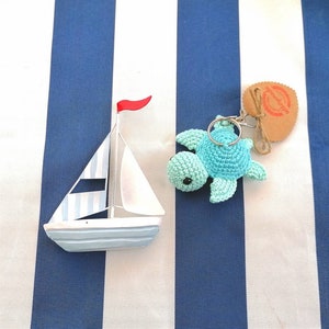 Könnte beinhalten: Ein blaues und weißes Segelboot mit einem roten Segel und einem blauen und türkisfarbenen gehäkelten Schildkröten-Schlüsselanhänger mit einem silbernen Schlüsselring. Die Gegenstände befinden sich auf einem weiß-blau gestreiften Hintergrund.