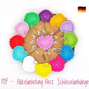 May include: A set of 10 crocheted heart-shaped keyrings in various colours, including pink, red, yellow, blue, green, and purple. Each keyring is attached to a silver key ring. The keyrings are arranged in a circle on a wooden surface. The text "PDF - Häkelanleitung Herz Schlüsselanhänger" is visible at the bottom of the image.