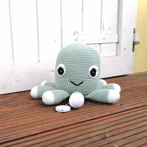 Könnte beinhalten: Ein grüner gehäkelter Oktopus mit weißen Akzenten und schwarzen Augen. Der Oktopus hat einen musikalischen Aufziehmechanismus auf der Unterseite.