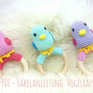 May include: Three colorful crochet bird rattles. Each bird is a different color: pink, teal, and purple. They have yellow beaks and accents, with wooden ring handles. The text "PDF - HÄKELANLEITUNG VOGELRASSEL" is visible.