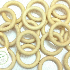 Könnte beinhalten: Eine Ansammlung hellfarbiger Holzringe, wahrscheinlich für Bastelarbeiten oder Babyspielzeug, liegt auf einer Oberfläche. Die Ringe sind glatt und haben eine natürliche Holzmaserung. Ein kleines Logo ist unten links zu sehen.
