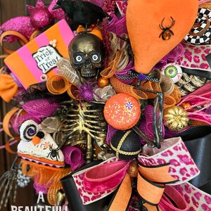 May include: A Halloween wreath with a pink, orange, and black color scheme. It features a skeleton, a skull, a crow, a spider, and a pumpkin. The wreath is decorated with ribbons, glitter, and other festive embellishments. The text "Trick or Treat" is visible on a pink and orange sign.
