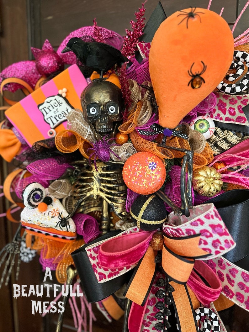 May include: A Halloween wreath with a pink, orange, and black color scheme. It features a skeleton, a skull, a crow, a spider, and a pumpkin. The wreath is decorated with ribbons, glitter, and other festive embellishments. The text "Trick or Treat" is visible on a pink and orange sign.