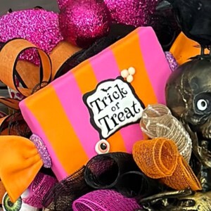May include: A pink and orange striped candy-shaped Halloween wreath with a "Trick or Treat" sign, black and orange mesh, a black crow, and a gold skeleton.