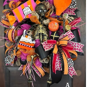 May include: A Halloween wreath featuring a skeleton, a black crow, a pink and orange candy box, a black and white checkerboard ribbon, a pink and black leopard print ribbon, and an orange balloon with a spider.