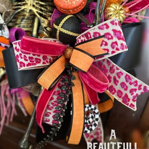 May include: A Halloween wreath with a black and white leopard print ribbon, a pink velvet ribbon, an orange ribbon, and a black and white checkered ribbon. The wreath also features a black and gold ornament, a skeleton, and a small pumpkin.
