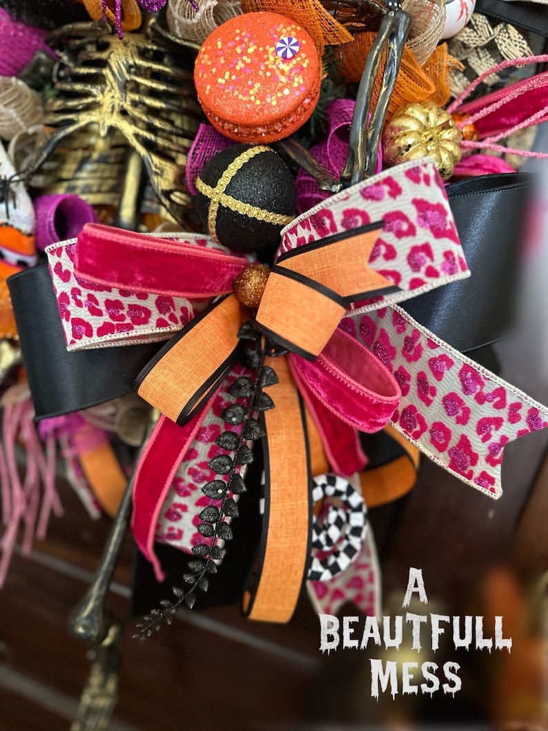 May include: A Halloween wreath with a black and white leopard print ribbon, a pink velvet ribbon, an orange ribbon, and a black and white checkered ribbon. The wreath also features a black and gold ornament, a skeleton, and a small pumpkin.