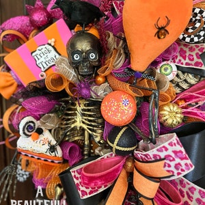 May include: A Halloween wreath with a pink, orange, and black color scheme. It features a skeleton, a black crow, a skull with googly eyes, a pumpkin, a spider, and a sign that says "Trick or Treat".