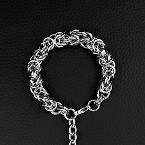 May include: A silver-toned chain bracelet featuring a complex Byzantine weave pattern. The bracelet includes a dangling extension chain that terminates in a small, circular charm embossed with a simple smiley face icon, creating a distinct look.