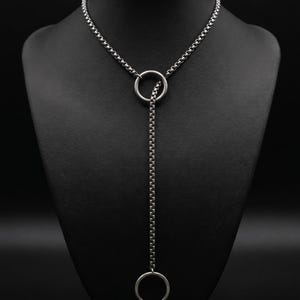 Op de afbeelding: Een zilverkleurige lariat-ketting met een box-chain ontwerp. De ketting loopt door een ronde ring in het midden en hangt naar beneden naar een tweede, kleinere ring aan het uiteinde. Een piepkleine, ronde bedel met een smiley is bevestigd.