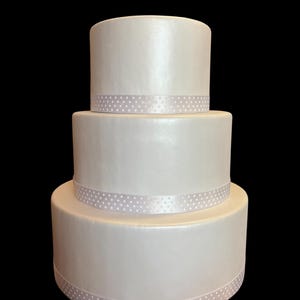 Könnte beinhalten: Ein dreistöckiger weißer Kuchen mit Perlmutt-Finish. Jede Etage ist mit einem hellrosa Band mit weißen Tupfen verziert. Der Kuchen steht vor einem schwarzen Hintergrund, der sein elegantes Design hervorhebt.
