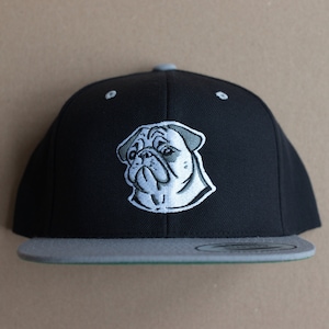 Könnte beinhalten: Eine schwarze und graue Baseballkappe mit einem gestickten Mops-Design. Die Kappe hat einen flachen Schirm und eine graue Unterseite.
