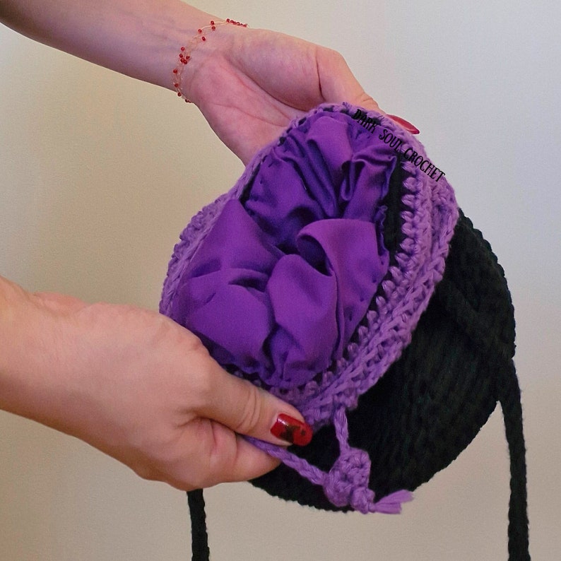 K&ouml;nnte beinhalten: Eine schwarze geh&auml;kelte M&uuml;tze mit einem violetten Futter und einem violetten Bommel. Die M&uuml;tze hat ein wei&szlig;es Etikett mit der Aufschrift "White Soul Crochet".