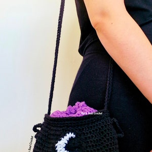K&ouml;nnte beinhalten: Eine schwarze geh&auml;kelte Kesseltasche mit einem wei&szlig;en Halbmond-Design. Die Tasche hat ein lila Futter und einen langen schwarzen Riemen.