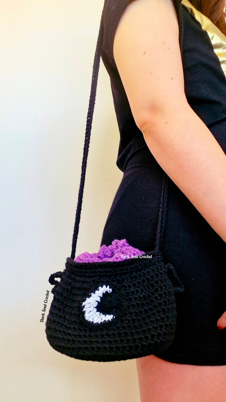 K&ouml;nnte beinhalten: Eine schwarze geh&auml;kelte Kesseltasche mit einem wei&szlig;en Halbmond-Design. Die Tasche hat ein lila Futter und einen langen schwarzen Riemen.