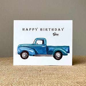 May include: A greeting card with a watercolor illustration of a blue vintage pickup truck. The card reads "HAPPY BIRTHDAY Son" in black text. The card is on a neutral surface.