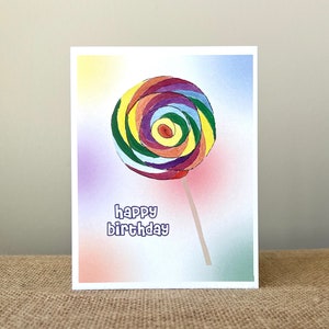 May include: A greeting card featuring a watercolor illustration of a colorful lollipop. The lollipop has a spiral design with shades of red, yellow, green, purple, and brown. The words "happy birthday" are written in a blue, bubble-style font.