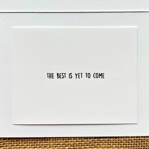 May include: A white greeting card with the phrase "THE BEST IS YET TO COME" printed in black. The card is set against a white background, with a textured, neutral-toned surface visible in the background.