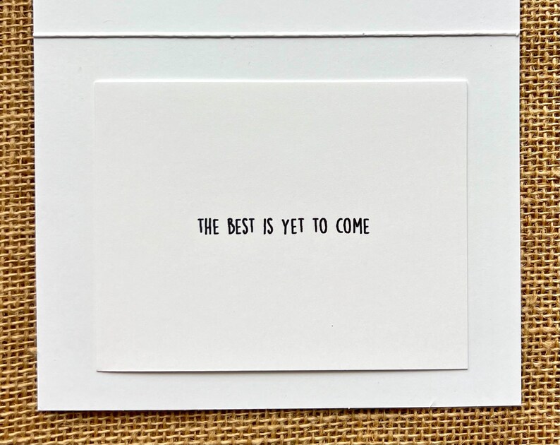 May include: A white greeting card with the phrase "THE BEST IS YET TO COME" printed in black. The card is set against a white background, with a textured, neutral-toned surface visible in the background.
