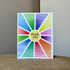 May include: A colorful thank you card with a sunburst design. The card features a white border and a central yellow circle with the words "thank you" in black. The sunburst is composed of watercolor segments in various colors, including blue, green, purple, and orange.
