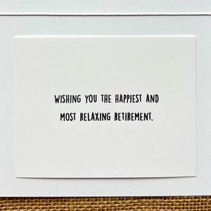 May include: A white greeting card with the message "WISHING YOU THE HAPPIEST AND MOST RELAXING RETIREMENT" in black text. The card is set against a textured, neutral-toned background.