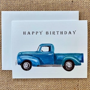 May include: A birthday card with a blue vintage truck illustration. The words "HAPPY BIRTHDAY" are printed in black above the truck. The card is white and comes with a white envelope.