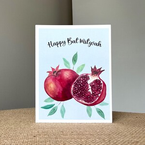 May include: A greeting card with a light blue background features a watercolor illustration of two pomegranates and green leaves. The words "Happy Bat Mitzvah" are written in black script.