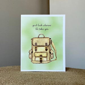 May include: Greeting card featuring a watercolor illustration of a tan backpack with brown straps and buckles. The card has the text "good luck wherever life takes you" on a light green background.