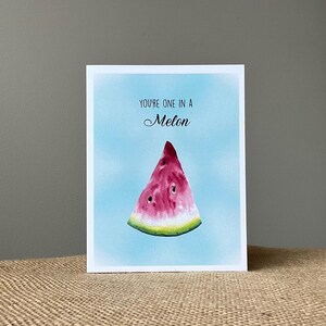 May include: A rectangular greeting card featuring a watercolor illustration of a single watermelon slice centered against a light blue background. The text "YOU'RE ONE IN A Melon" is printed in black ink above the fruit, with "Melon" in script.