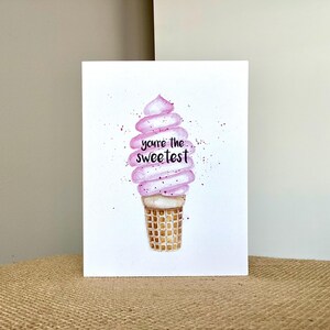 May include: A greeting card featuring a watercolor illustration of a soft-serve ice cream cone. The ice cream is swirled pink with the words "you're the sweetest" written in black. The cone is a waffle pattern.