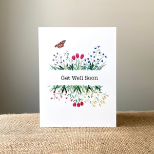 May include: A white greeting card with a watercolor floral design and the text "Get Well Soon." The card features a butterfly and colorful flowers in shades of red, blue, and yellow.