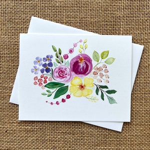 May include: A set of blank greeting cards with a watercolor floral design. The artwork features purple, pink, and yellow flowers with green leaves and red berries. The cards are white, and the background is a neutral tan.