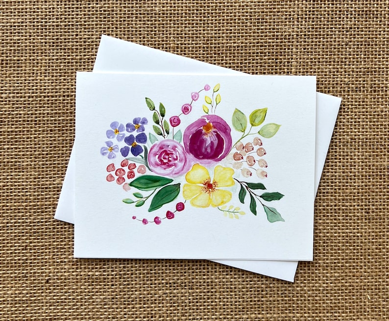 May include: A set of blank greeting cards with a watercolor floral design. The artwork features purple, pink, and yellow flowers with green leaves and red berries. The cards are white, and the background is a neutral tan.