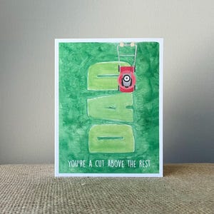 May include: A greeting card with a green background and the word "DAD" cut out of the grass. A red lawnmower is positioned above the "A". The text "YOU'RE A CUT ABOVE THE REST" is at the bottom.