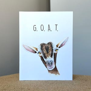 May include: A white greeting card featuring a colored pencil illustration of a brown and white goat head centered below the text G. O. A. T. printed in a simple black sans-serif font. The goat has yellow eyes and detailed fur texture.