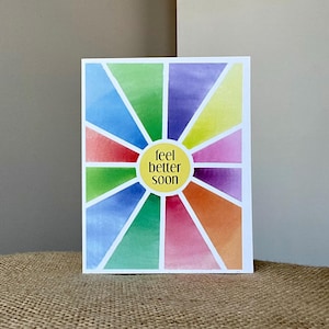 May include: A rectangular greeting card with a colorful watercolor design. The card features a sunburst pattern with segments of blue, green, yellow, purple, orange, and pink. A yellow circle in the center reads "feel better soon."
