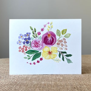 May include: A white greeting card featuring a watercolor floral design. The arrangement includes purple, pink, and yellow flowers with green leaves and red berries. The artwork is painted in a loose, expressive style.