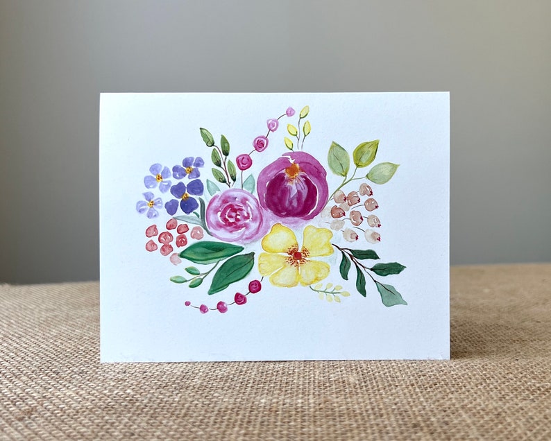 May include: A white greeting card featuring a watercolor floral design. The arrangement includes purple, pink, and yellow flowers with green leaves and red berries. The artwork is painted in a loose, expressive style.