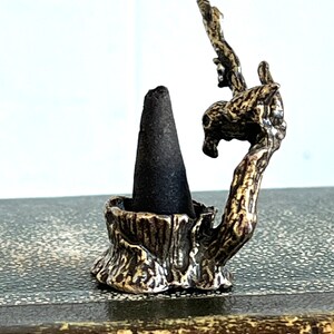May include: A small, gold-toned metal incense holder shaped like a tree branch with a cone-shaped incense burner. The holder is sitting on a brown, textured surface.