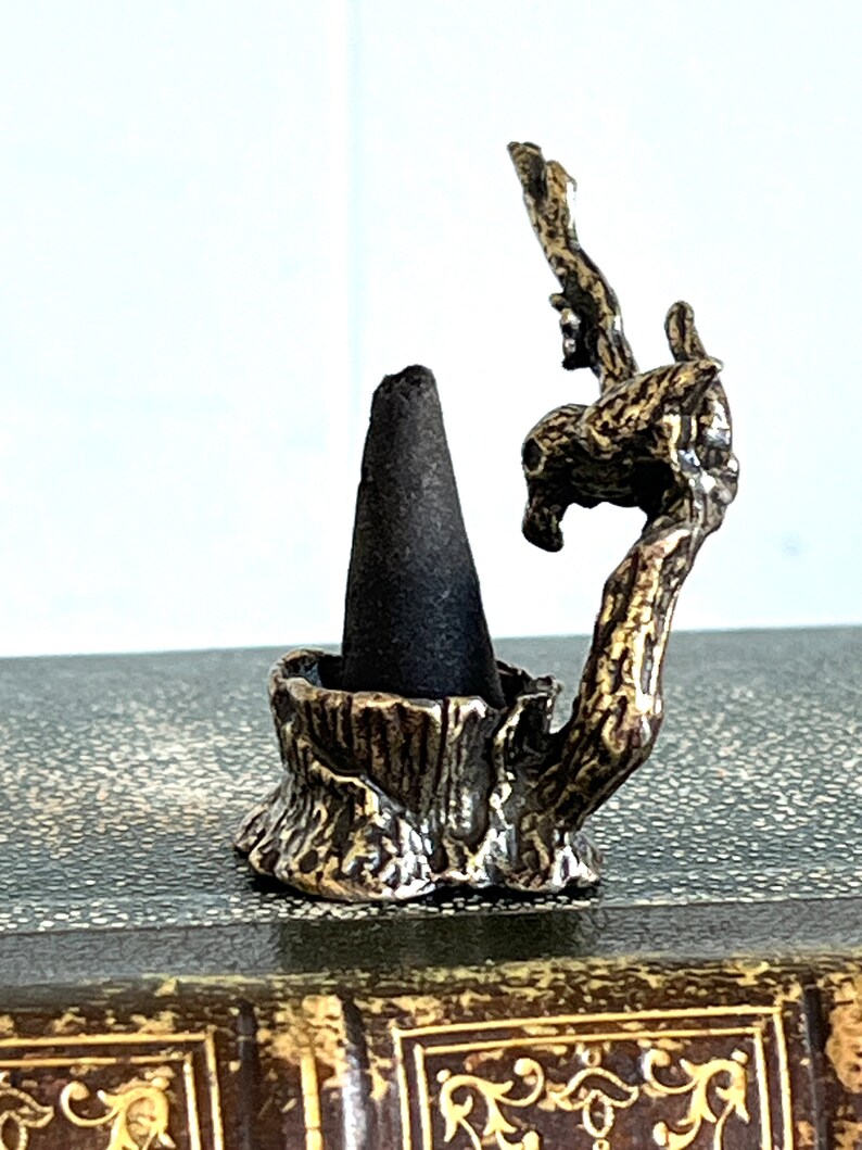 May include: A small, gold-toned metal incense holder shaped like a tree branch with a cone-shaped incense burner. The holder is sitting on a brown, textured surface.