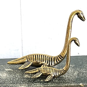 May include: Two brass-colored sculptures of plesiosaurs, one large and one small, with long necks and detailed bodies. The sculptures are positioned on a textured surface, showcasing the intricate design and metallic finish.