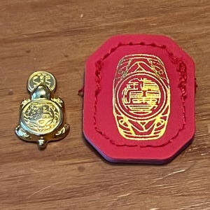 May include: A gold-colored turtle figurine and a red, octagonal-shaped item with gold-colored Chinese characters. The turtle has a coin on its head. The red item has a gold design and a stitched border.