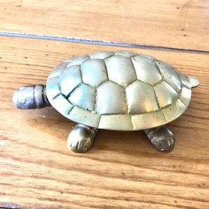 May include: A gold-colored metal turtle figurine with a detailed shell and four legs. The turtle is sitting on a wooden surface.