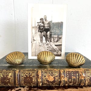 May include: Three gold-colored seashell-shaped decorative objects are arranged in front of a vintage black and white photograph of a couple. The photograph is propped up on a stack of antique books with gold lettering.