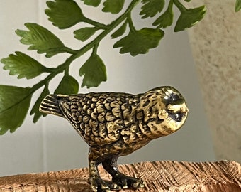 May include: A small, gold-colored metal owl figurine with detailed feathers and large eyes. The owl is perched on a piece of weathered wood.